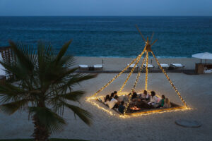 picture of people sitting in a open cafe on a beach decorated with yellow lights with sea in front