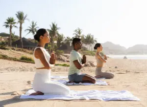 Three people practicing yoga on mats on a sandy beach with palm trees and ocean in the background.