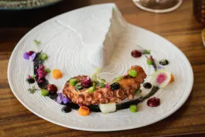 Pulpo gourmet servido con verduras de colores, flores comestibles y salsa en un plato blanco texturizado.